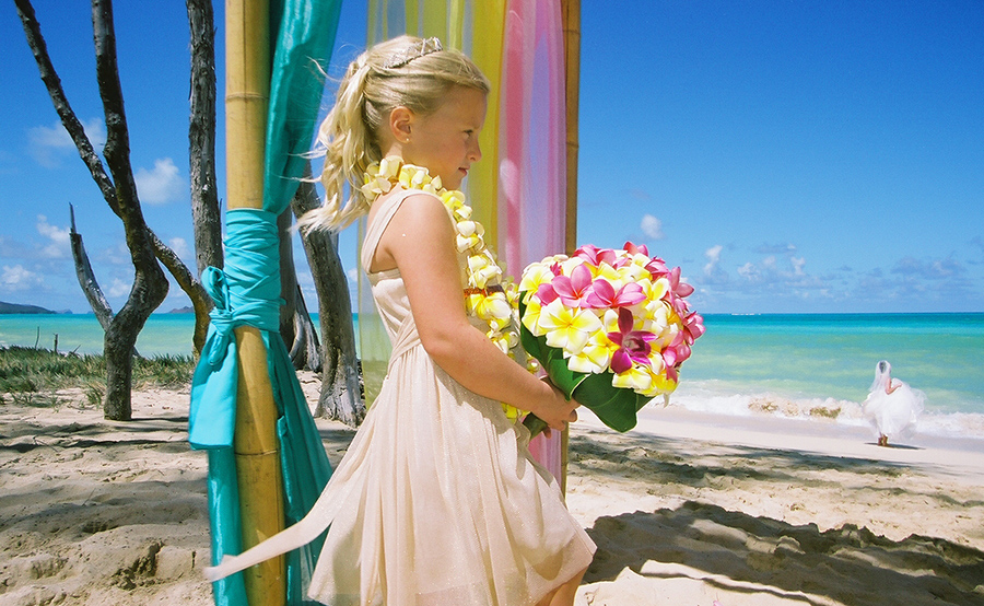 Girl holding a colorful plumeria bouquet beside a turquoise draped arch on the beach